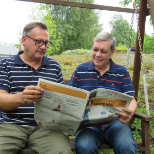 Två män sitter i en trädgårdsgunga och tittar i en papperstidning, Hangötidningen. Sommar, utomhus.