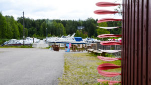 Vid hamnen vilar både gästbåtar och kajakpaddlar. Bakom ett rött skjul till höger sticker en massa paddlar ut. Längre fram syns stranden med flera båtar.