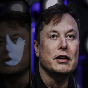 Elon Musk näkyy hartioista ylöspäin ja hän katsoo yläviistoon. Muskin vasemmalla puolella näkyy näyttö, jossa on Twitterin logo ja jonka pinnasta heijastuvat Muskin kasvot.