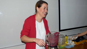 Rektor Petra Blomqvist lagar smoothies så att hennes abiturienter ska ha kraft att åka ut på stan på lastbilsflak. En kvinna i röd tröja och vit skjorta fixar smoothie med hjälp av en mixer/maskin för smoothies. Hon är i ett klassrum och ser glad ut.