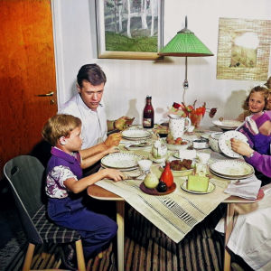 familj från 1950 talet som sitter vid ett köksbord och äter middag. Familjen ser glad och stämningen är trivsam. Modern har ett förkläde på sig. Man ser att det är hon som fixat middagen. 