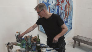 Taidemaalari Mikko Sakala ateljeessaan Taidekeskus Antareksessa Sippolassa maalipurkkien äärellä, taustalla hänen sini-puna-sävyinen teoksensa.