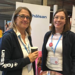 Katarina Fagerström och Saija Westerlund-Cook två leende kvinnor med glasögon står vid en skylt med Folkhälsan. Den ena har en munktröja där det står Folkhälsan och en papperskaffekopp i handen