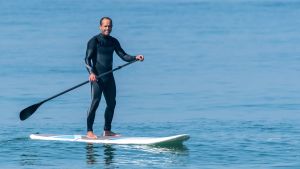 SUP, eller Stand Up Paddling som träningsformen heter. En glad man i mörk våtdräkt på en paddelbräda i ett blått hav. Han har en paddel i handen. Solen skiner. Sportig stämning.