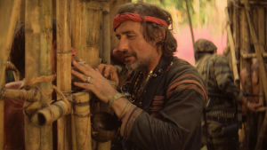 Dennis Hopper spelar en hänförd och upphetsad fotojournalist. Fotojournalisten (Dennis Hopper) står vid ett bambustängsel.