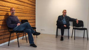 Maksimainen och Martin planerar var tavlorna ska hänga. Två män sitter på var sin stol i ett rum med en vit vägg och en stockvägg.