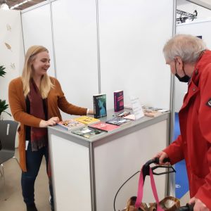 Lotta Westerlund, en dam med ljust lång hår, står vi en montor på bokmässan och diskuterar böcker med en man i röd jacka.