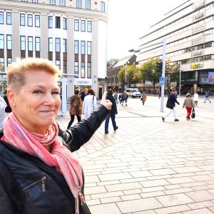En kvinna med kort ljust hår, svart läderjacka och en rödvit halsduk pekar mot en stor vit byggnad i en korsning, där folk går över gatan.