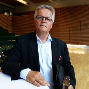 Torbjörn Kevin har skrivit en SFP-historik. Tidigare chefredaktör Torbjörn Kevin står vid ett bord i ett auditorium med en mapp under armen.