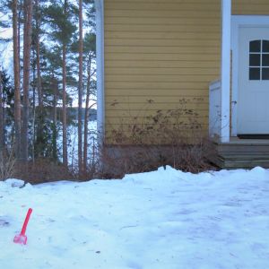 En röd plastspade (för lek i sandlådan) i snön utanför ett gult trähus. Hav i bakgrunden eller egentligen is. Vinter, snö och is.