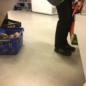 Kunderna släpps in i små grupper. Var och en får gå från disk till disk för att få eller själv plocka med sig de livsmedel som finns den dagen. Ett par fötter i svarta joggingskor, matkassar som personen håller i, bakom personen syns en korg med potatis.