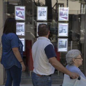 Turismen har bidragit till att bostadspriserna i Lissabon skjutit i höjden. En medelålderspar och en äldre dam i rullstol tittar på ett skyltfönster med bostadsannonser i Lissabon.