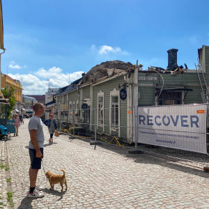 En man håller en liten hund i ett koppel. Han står i Gamla stan i Borgå och ser upp mot ett hus vars tak brunnit. Det är en varm sommardag och solen skiner.