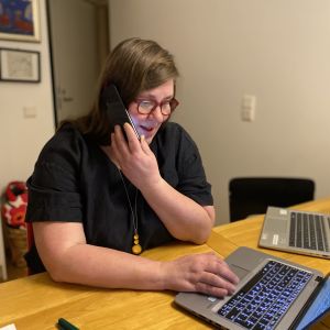 Kirsi Lehtimäki istuu työpöydän ääressä ja pitää toisella kädellä puhelinta korvalla ja toisella kädellä käyttää kannettavaa tietokonetta.