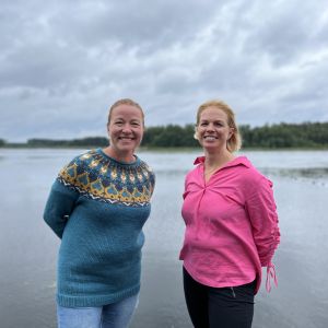 Kvinna i blått och kvinna i rosa ler motkameran framför hav.