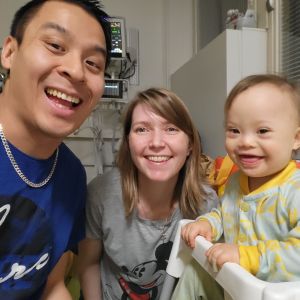 En familj på tre personer i sjukhusmiljö tittar alla tre in i kameran och ler. Familjen består av en pappa, en mamma och en bebis som har downs syndrom.