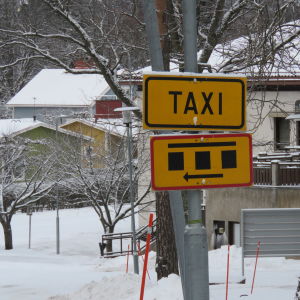 En taxiskylt i gult med svart text och en skylt som visar marlerade parkeringsrutor på en stolpe utanför Raseborgs sjukhus i Ekenäs. Man ser inte sjukhuset men nog en del småhus. Vinter och snö. 