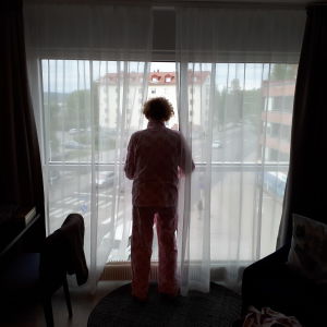 nainen katsoo ulos hotellihuoneen isosta ikkunasta valoa vasten