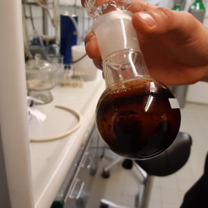 brun olja i runt glaskärl i laboratorium. En hand håller kärlet