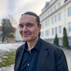 Personporträtt på man som ler och tittar in i kameran. Han är klädd är mörkblå skjorta och kavaj, i bakgrunden våningshus med ljusgul rappning.