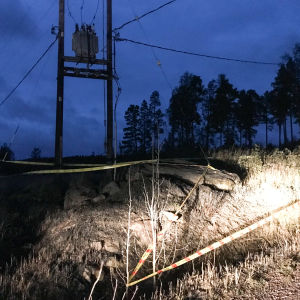 Transformatorn måste bytas ut. Det luktar rök och det finns aska på marken. Området har spärrats av med plastband. På bilden syns en transformator uppe mellan några stolpar. Framför transformatorn har man satt ut varningsband.