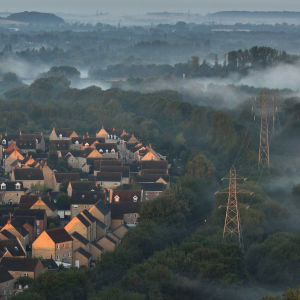 Landskapsbild över en liten engelsk by dränkt i dimma vid en grön skog. I utkanten av byn, till höger i bild, tornar sig stora kraftledningar upp ur dimman. 