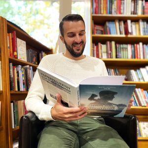En man i vit tröja sitter omgiven av bokhyllor med en bok i sin hand.
