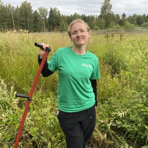 En ung kvinna med blont hår står på en åker med en lie i handen.