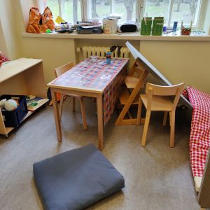 Ett rum med bara väggar och ett bord och några stolar i barnstorlek. Diverse påsar, pappershögar och burkar belamrar fönsterbrädet. 