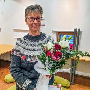 En kvinna i grå ylletröja och glasögon ler mot kameran och håller i en blombukett
