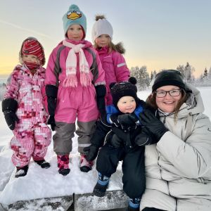 Neljä lasta ja yksi aikuinen poseeraavat kameralle talvisissa ja lumisissa maisemissa. 