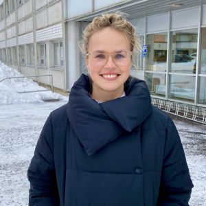 En glad Malin Olkkola står utomhus i blå vinterjacka, glasögon och uppsatt hår.