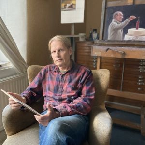 Taidemaalari Kaj Stenvall istuu nojatuolissa työtilassaan kädessään yksi teoksistaan. 