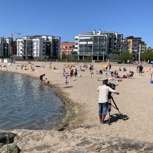 Här filmar Yle Tv-Nytt på Solvikens badstrand på midsommarafton. Hundratals personer njuter av det varma vädret och många simmar också.