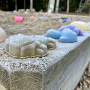 Sinisiä, punaisia, keltaisia ja violetteja hiekkakkumuotteja hiekkalaatikon reunalla. Muottien päällä on kuuraa.