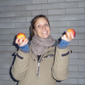 En skrattande och spexande Petra Österberg med ett rött äpple i var hand. I övrigt grå bakgrund och rätt mörka färger.