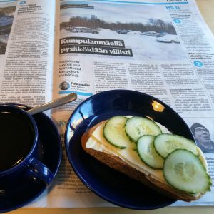 För en närståendevårdare kan dagens avkoppling vara att få läsa dagstidningen och hinna dricka en kopp kaffe. Kahvikuppi ja juusto-kurkkuvoileipä, siniset Teema-astiat sanomalehdellä