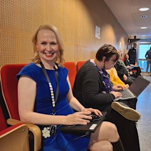 Nainen hymyilee leveästi kokoussalin taaimmaisella penkkirivillä. Vieressä muita osallistujia.