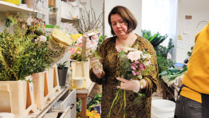 Saara Manninen och hennes syster driver två blombutiker tillsammans. Deras personal består av tre personer. En kvinna står bakom disken i en blombutik och plockar ihop en blombukett.