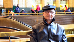 Timo Mäcklin minns barndomens valdagar då pappa hade kostym och man fick gräddtårta. Timo Mäcklin sitter på en bänk i Helsingfors Centralstation.