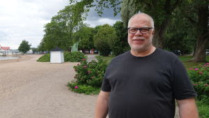 Magnus Boberg lär ut datorkunskaper. Magnus Boberg står på en strand med buskar och träd i bakgrunden. Han ler och har glasögon.