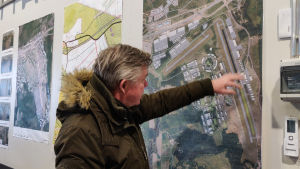 Så här kan flygplatsen komma att se ut om flera år. Korjula visar på ritningen som finns med i delgeneralplanen. Hangaren vi befinner oss i ligger ungefär vid Korjulas hand. En man i dunjacka pekar på en kartskiss som finns på en vägg.