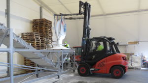 Munskydd är förstås bra också mot små dammpartiklar i luften, men det är nog coronaepidemin som har lett till att alla bär munskydd när de jobbar inne i hallen. En orange-röd gaffeltruck tömmer ut en stor säck med salt längs ett band som löper vidare mot en stor tankbil (bilden syns inte). Inne i en stor hall.