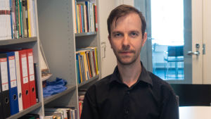 Johannes Kananen jobbar vid Helsingfors universitet. Johannes Kananen i en svart skjorta stående bredvid en bokhylla.