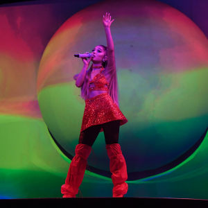 Laulaja Ariana Grande seisoo värikkään screenin edessä lavalla toinen käsi ilmaan nostettuna. Toinen käsi pitelee mikkiä suun edessä. Päällään hänellä on punaiset korkeat saappaat ja punainen paljettiasu.