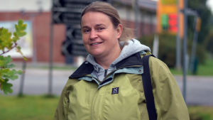 Susse Ekström har varit aktiv i kommunalpolitiken i Sjundeå i 20 år. Hon har varit med om både inflyttningstrenden och utflyttningsvågen. En kvinna med grön jacka står utomhus, framför en market. I bakgrunden ser man också vägskyltar. Hon tittar in kameran.