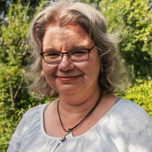 Vaaleatukkainen, vaaleaan pellavapaitaan pukeutunut nainen puutarhassa.