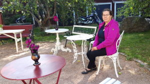 Anette Kanteh brukar guida grupper i Neristan i Karleby. Porträttfoto av Anette Kanteh som sitter vid ett gammaldags trädgårdsbord i en trädgård.