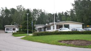 Hälsocentralen har haft problem med inomhusluften. Hälsocentralbyggnad på gräsplan vid asvalterad uppfart. Parkering utanför.
