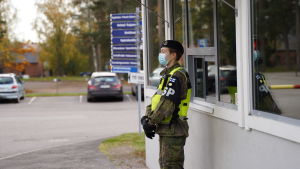 Den som har vakt vid porten kommer i kontakt med många personer och måste därför ha munskydd. Också i situationer där man inte kan hålla säkerhetsavstånd rekommenderas munskydd. En person i unifrom står med munskydd på sig vid ett vaktbås, I bakgrunden ett trafikljus som visar rött. Det är höst för löven är gula.